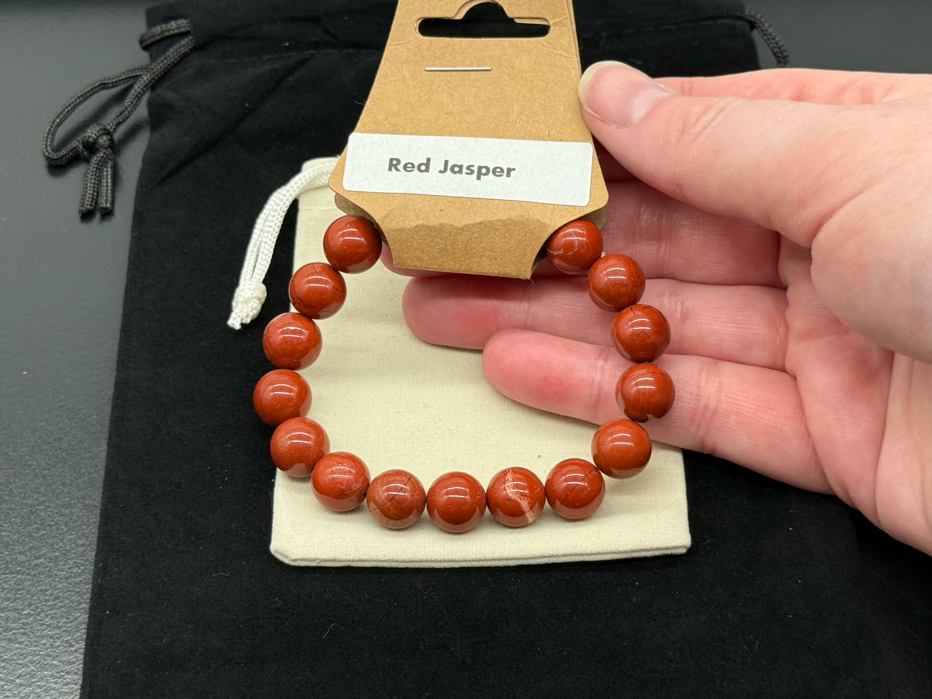 A polished red jasper stretch bead bracelet held in an open hand above a black drawstring pouch and a Red Jasper hang tag