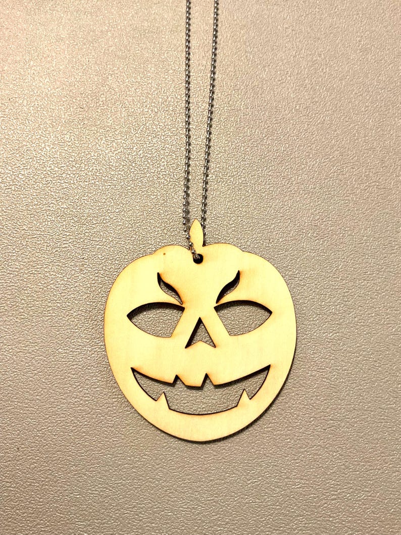 Close-up of a laser cut wood jack-o'-lantern pendant necklace showing the clean cut edges of the carved pumpkin face and the stainless steel ball chain — Robert Anthony Art laser cut Halloween jewelry
