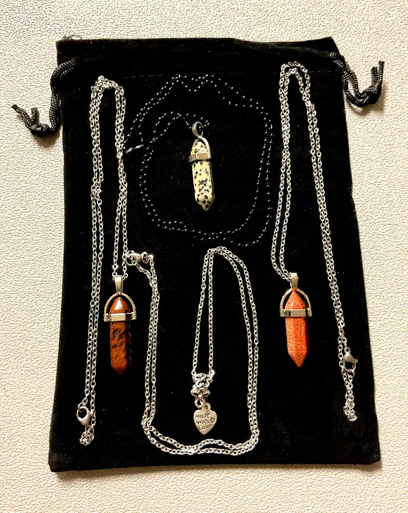 Flatlay of natural stone point pendants — dalmatian jasper, red jasper, and goldstone — with a 'made with love' charm on a black velvet pouch, photographed in the Robert Anthony Art home studio
