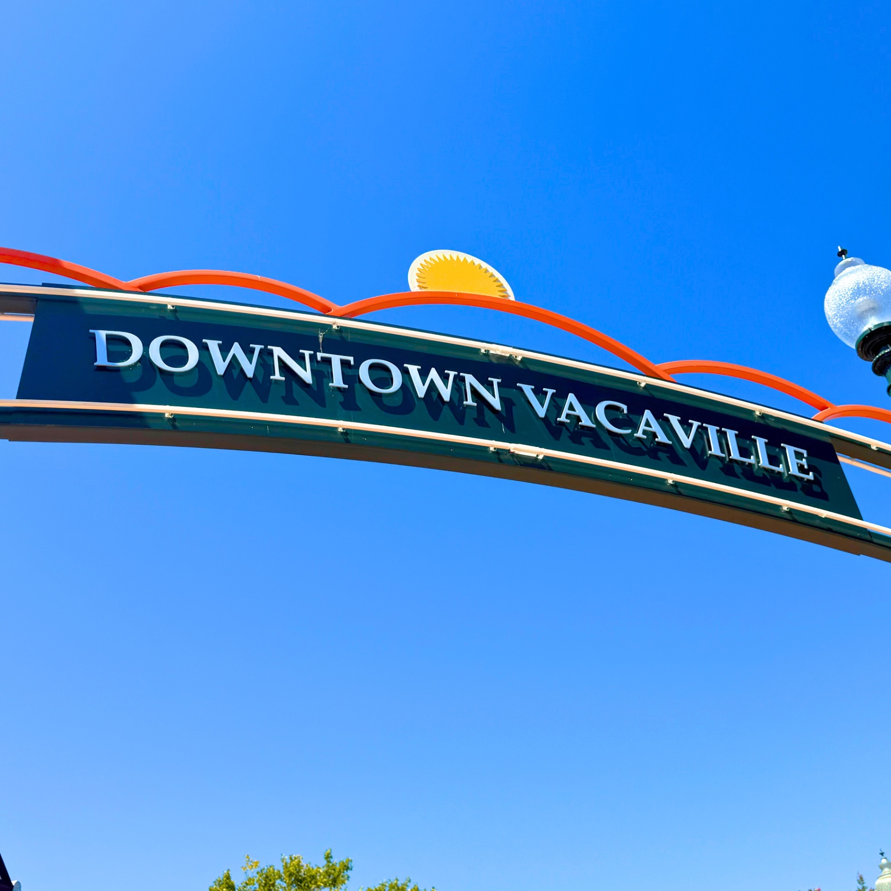 The Downtown Vacaville arch sign in Vacaville, California — green sign with white lettering and orange arches against a clear blue sky, photographed by Robert and Angela of Robert Anthony Art