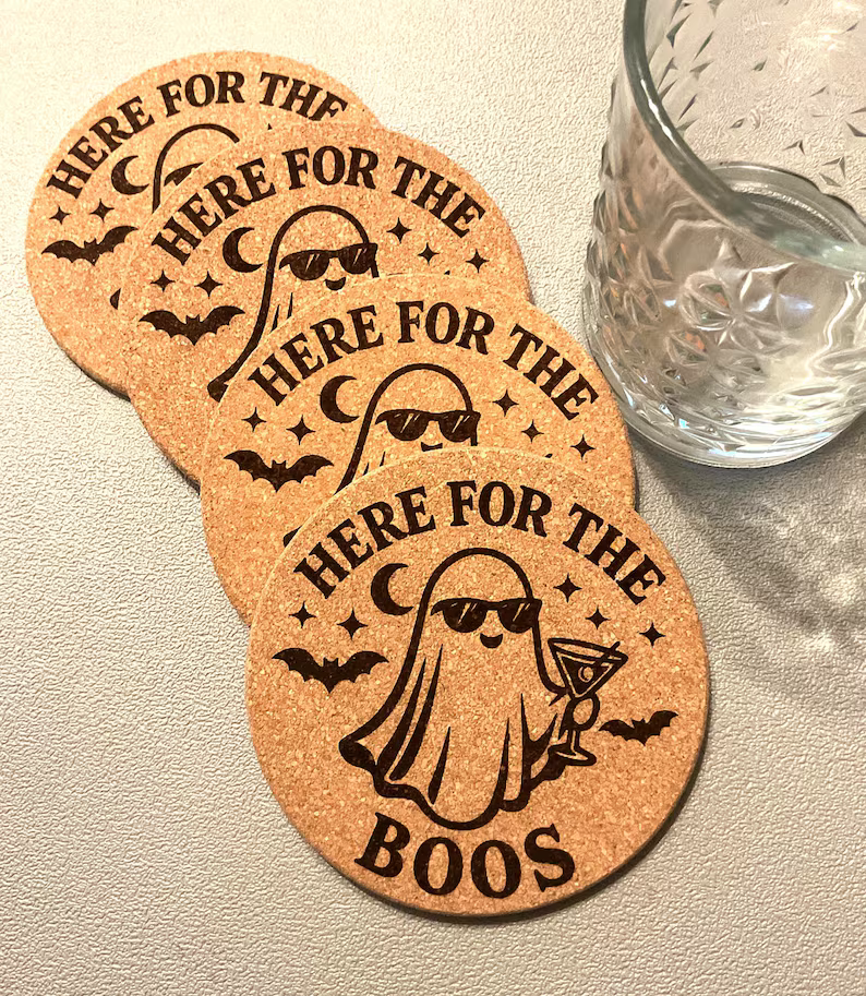 Set of four Halloween 'Here for the Boos' laser engraved cork coasters fanned out beside a cut crystal glass, each engraved with a ghost in sunglasses holding a martini, bats, stars and a crescent moon — custom laser engraving by Robert Anthony Art in Vacaville, California