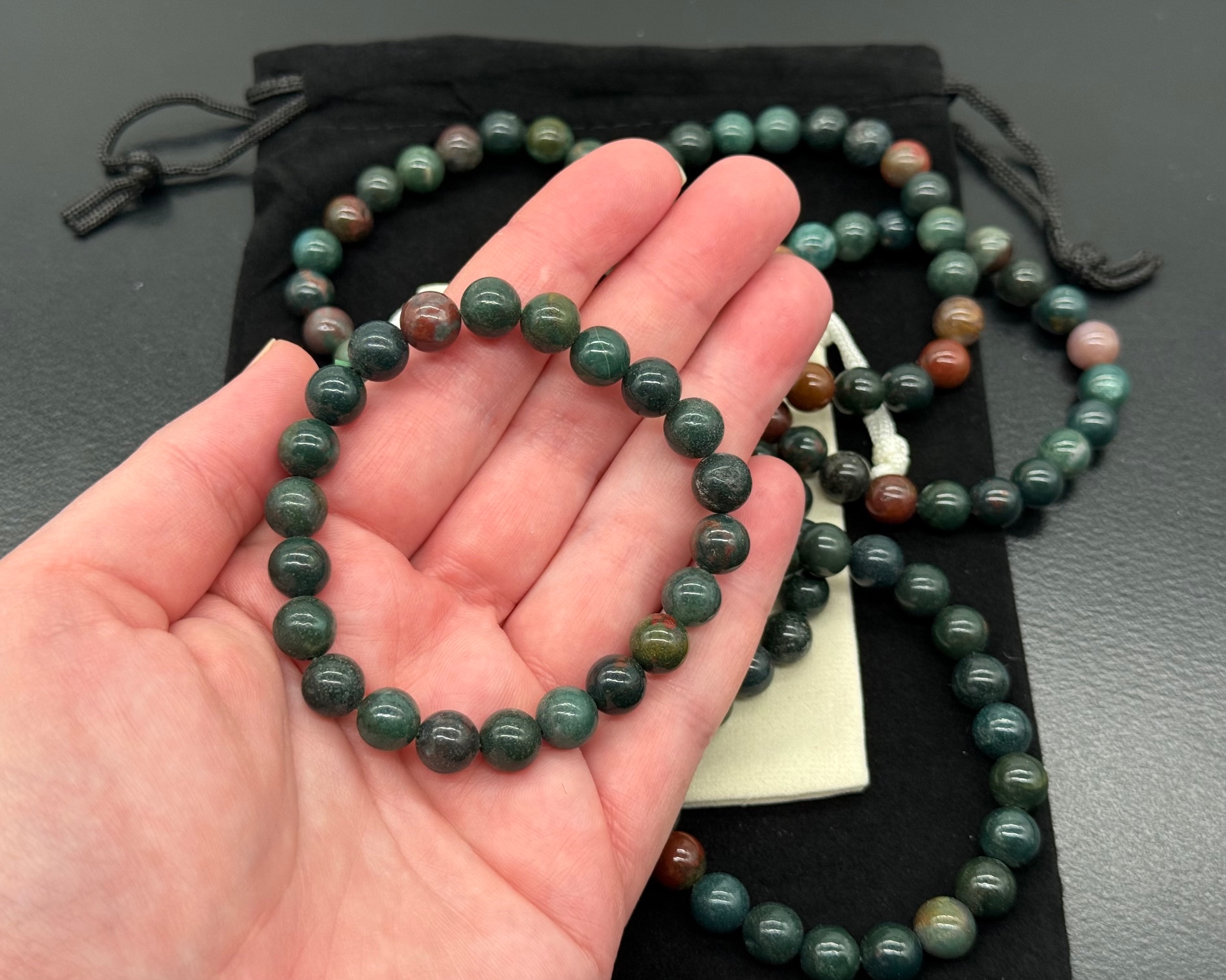 A dark green bloodstone stretch bead bracelet with rust-red flecks resting in an open hand, with a matching necklace and black pouch in the background