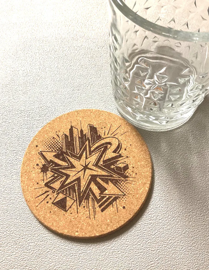 A single laser engraved cork coaster with a detailed cityscape and starburst design beside a cut crystal glass — the kind of small-format work a compact desktop diode laser engraver produces