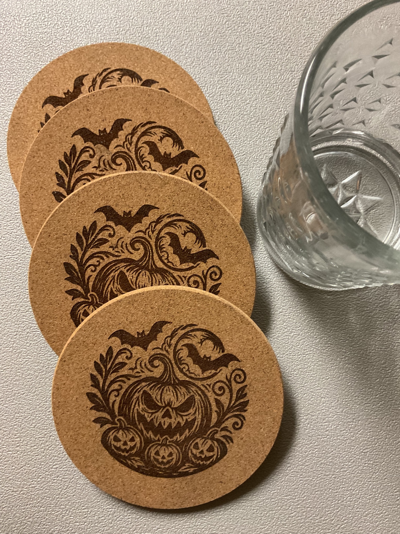 Set of four Halloween cork coasters laser-etched with jack-o'-lantern and bat designs, next to a crystal rocks glass