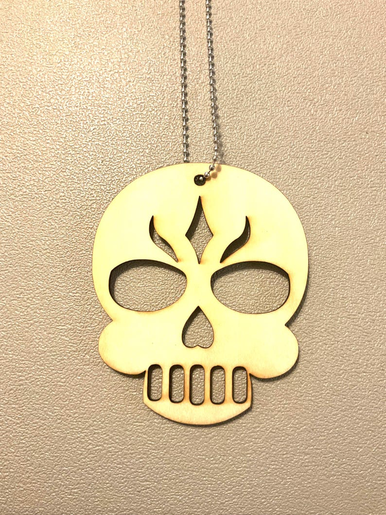 Laser cut birch skull pendant with flame-shaped brow detail and teeth cutouts hanging from a stainless ball chain against a warm textured wall