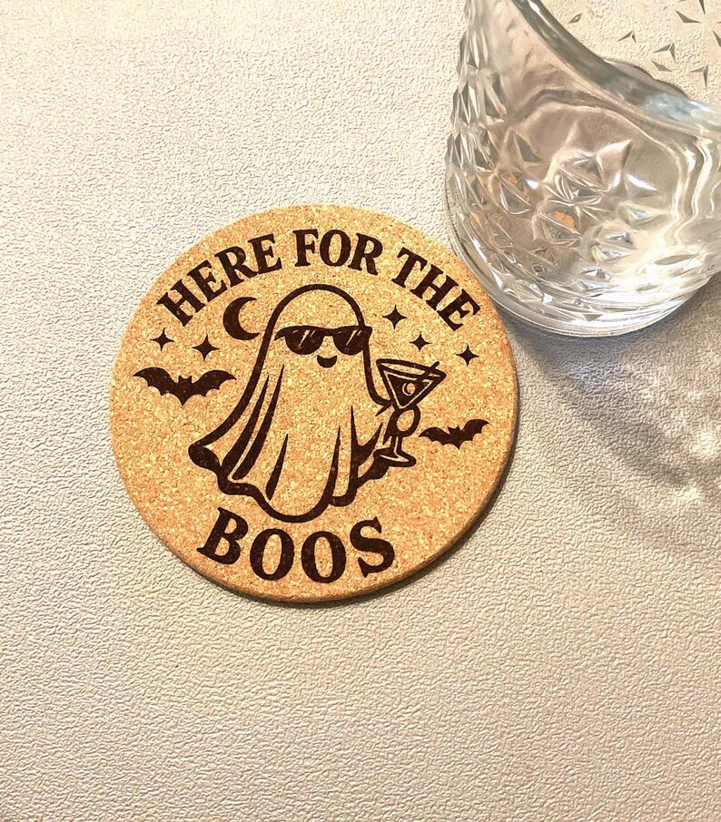 Single Halloween 'Here for the Boos' laser engraved cork coaster showing a cocktail-drinking ghost in sunglasses surrounded by bats and stars, photographed next to a cut crystal glass — Robert Anthony Art custom laser engraving