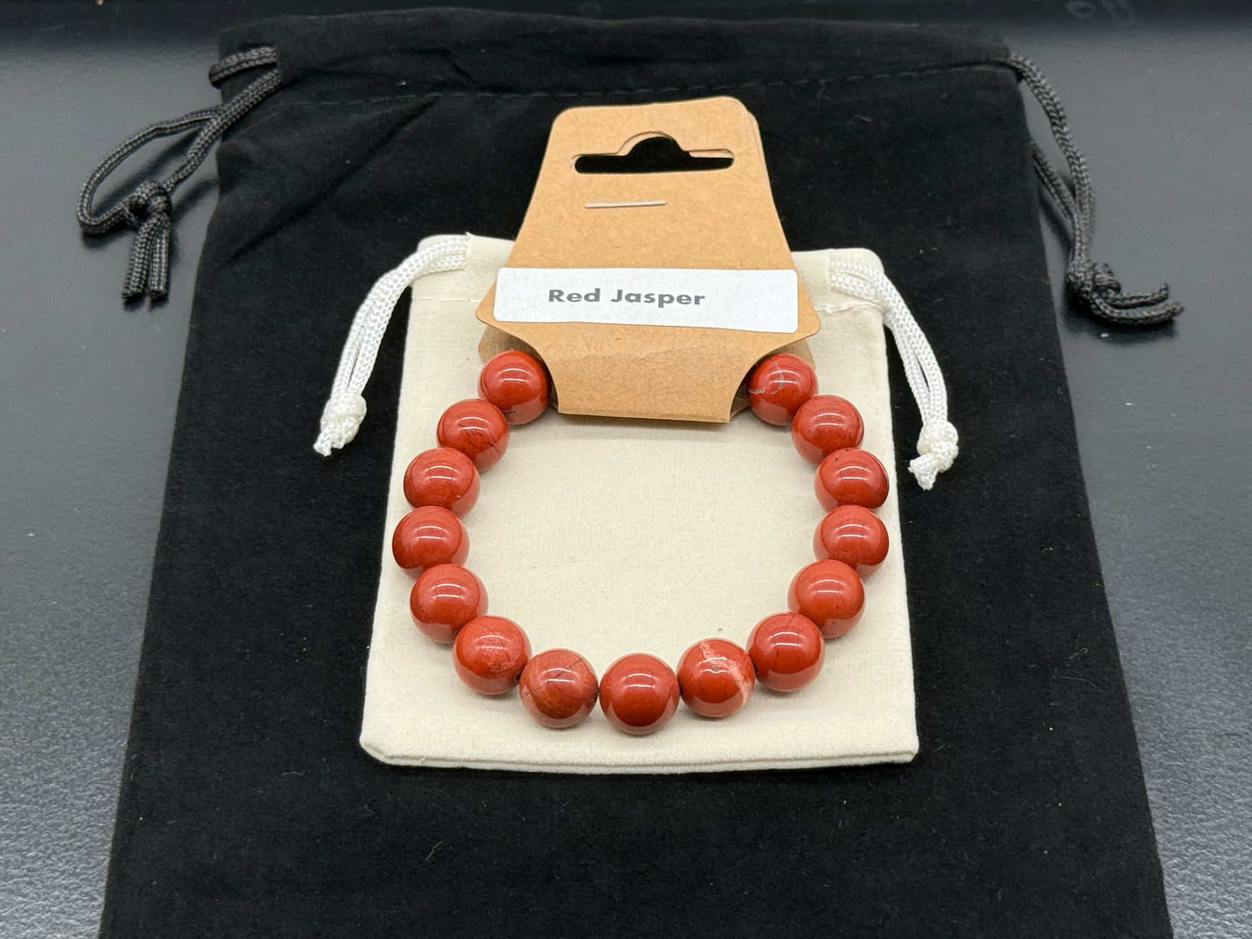 Red jasper bead bracelet with hangtag on a cream pouch, photographed in-studio by Robert and Angela
