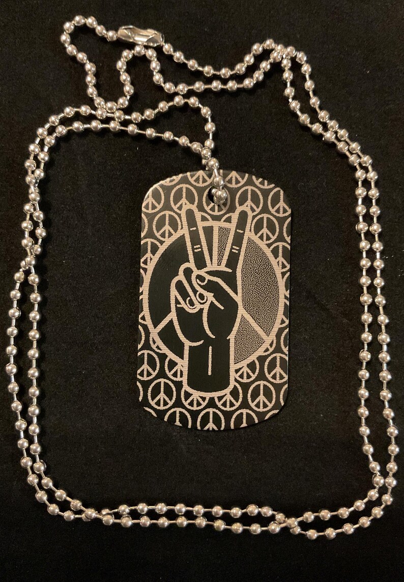 Custom laser engraved peace hand sign aluminum dog tag on a classic stainless steel ball chain, coiled in a spiral against a black velvet surface — hand made by Robert Anthony Art in Vacaville, California