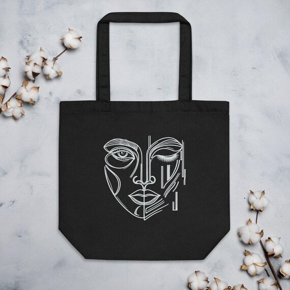 Black eco cotton tote bag printed with a white One-Eyed Muse hand-drawn face design, styled with cotton branches