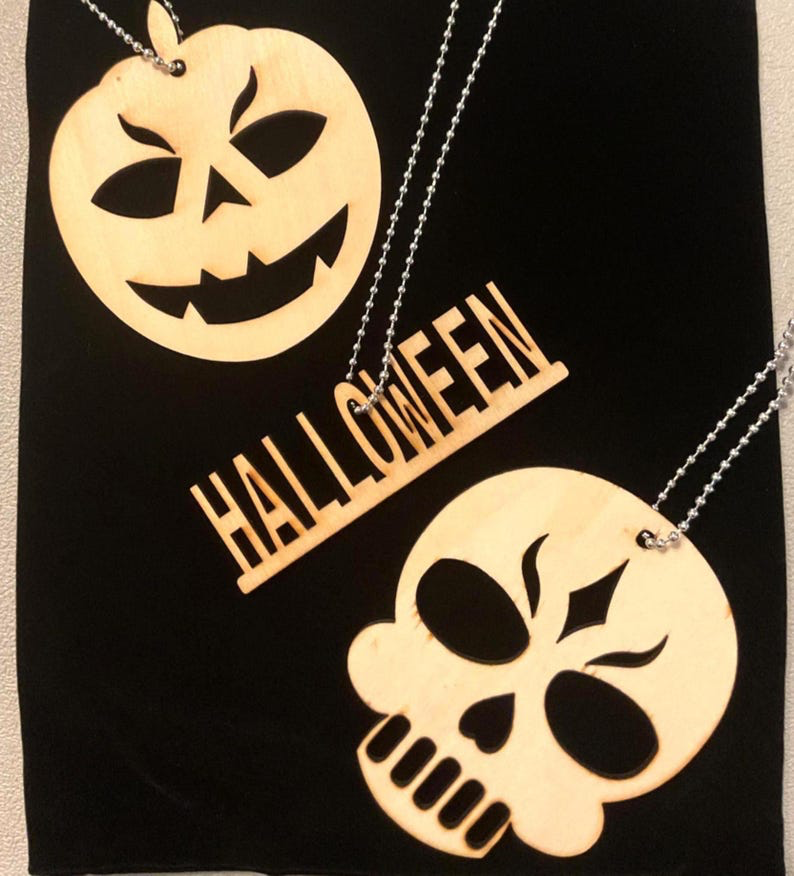 Three laser cut birch Halloween pendants on ball chains — a jack-o-lantern face, a HALLOWEEN nameplate, and a skull — arranged on black fabric