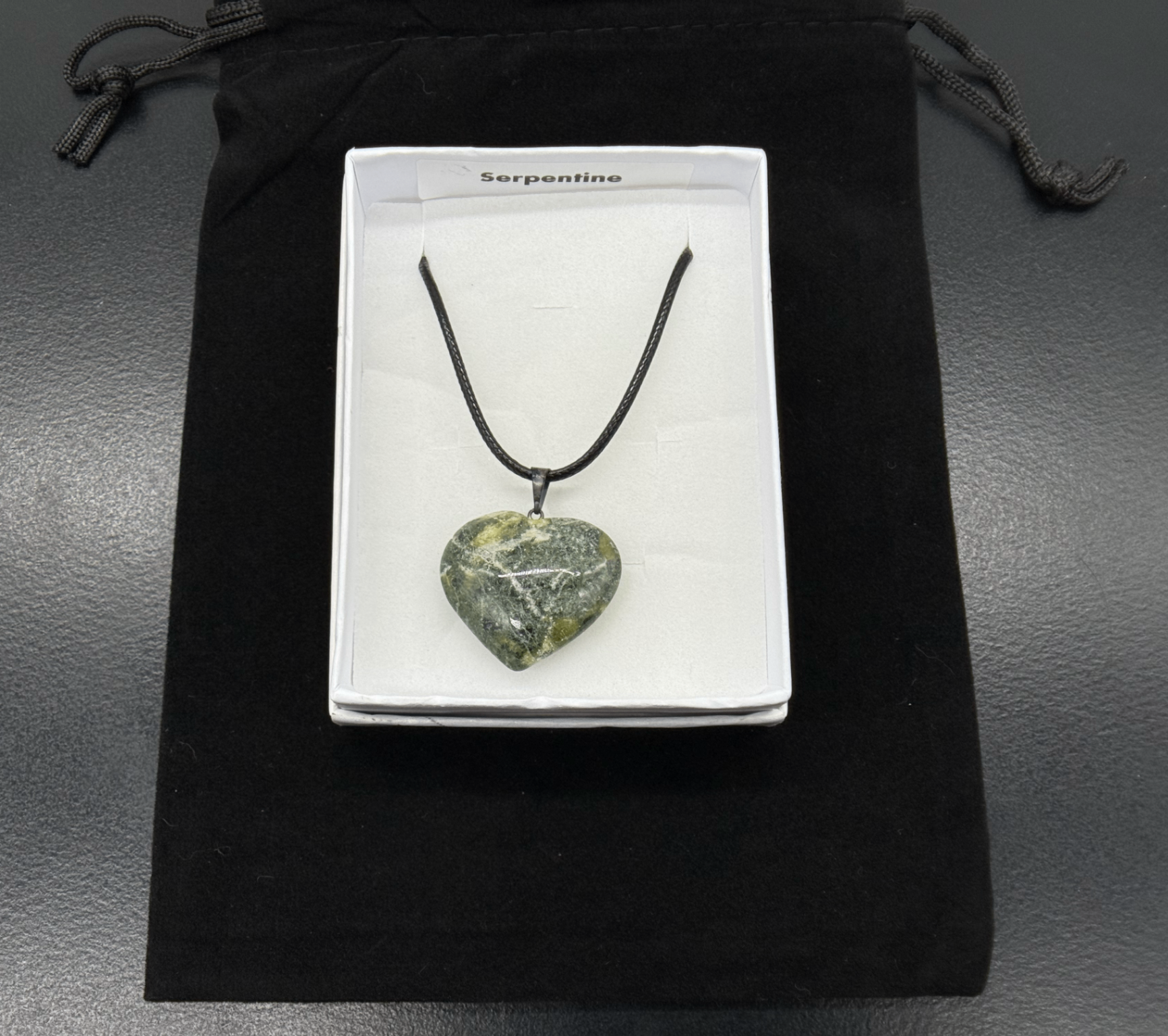 Polished serpentine heart pendant in a white gift box resting on a black velvet pouch, showing the mossy green and cream veining