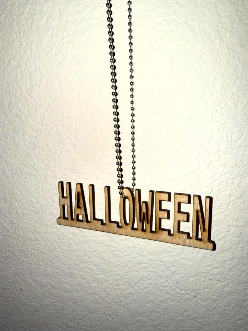Laser cut wood HALLOWEEN word pendant necklace in bold block letters on a stainless steel ball chain, hanging against a white wall — handmade laser cut jewelry by Robert Anthony Art