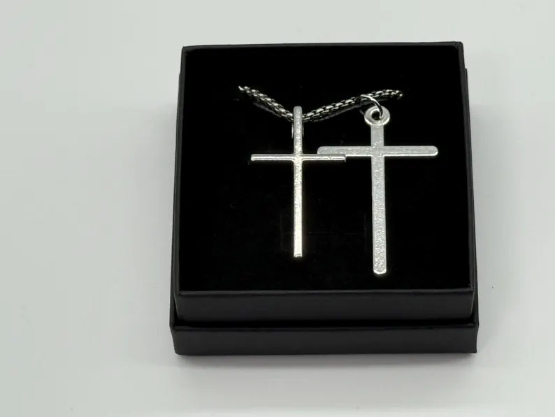 Two silver cross pendants — one brushed, one polished — resting side by side in a black jewelry box