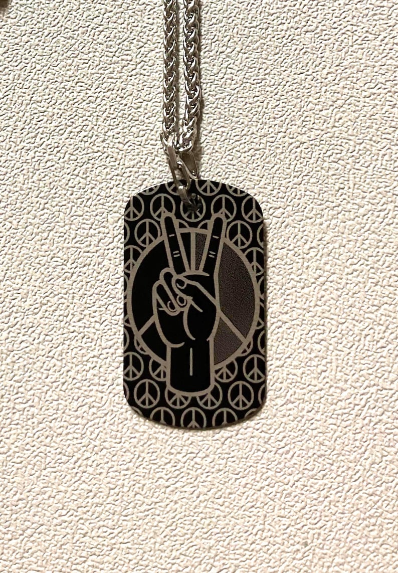 Custom laser engraved aluminum dog tag featuring a raised peace hand sign inside a peace symbol circle, set against a repeating pattern of small peace signs — hand made by Robert Anthony Art