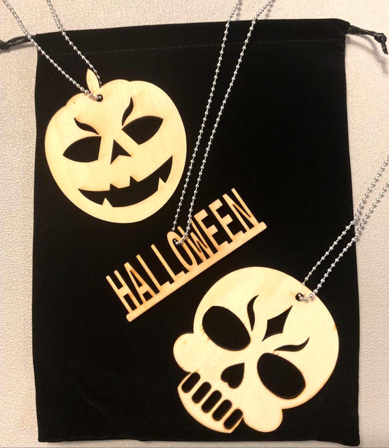 Complete three-piece Halloween wood necklace set displayed on a black velvet drawstring gift pouch — pumpkin, HALLOWEEN word bar and skull pendants on ball chains, laser cut by Robert Anthony Art