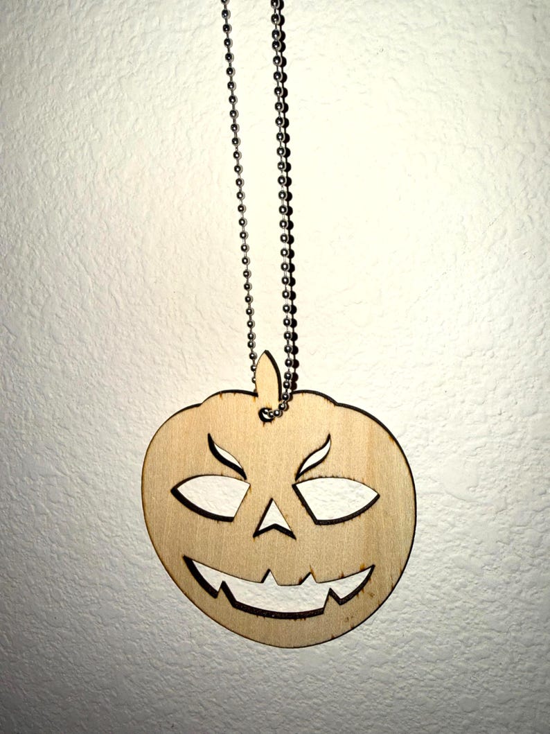 Laser cut birch wood jack-o'-lantern pendant necklace with a carved grinning face on a stainless steel ball chain, hanging against a white textured wall — Robert Anthony Art laser cutting